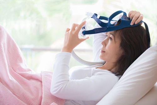 Woman in white clothes laying with knees up in bed hands holding and wearing CPAP mask .